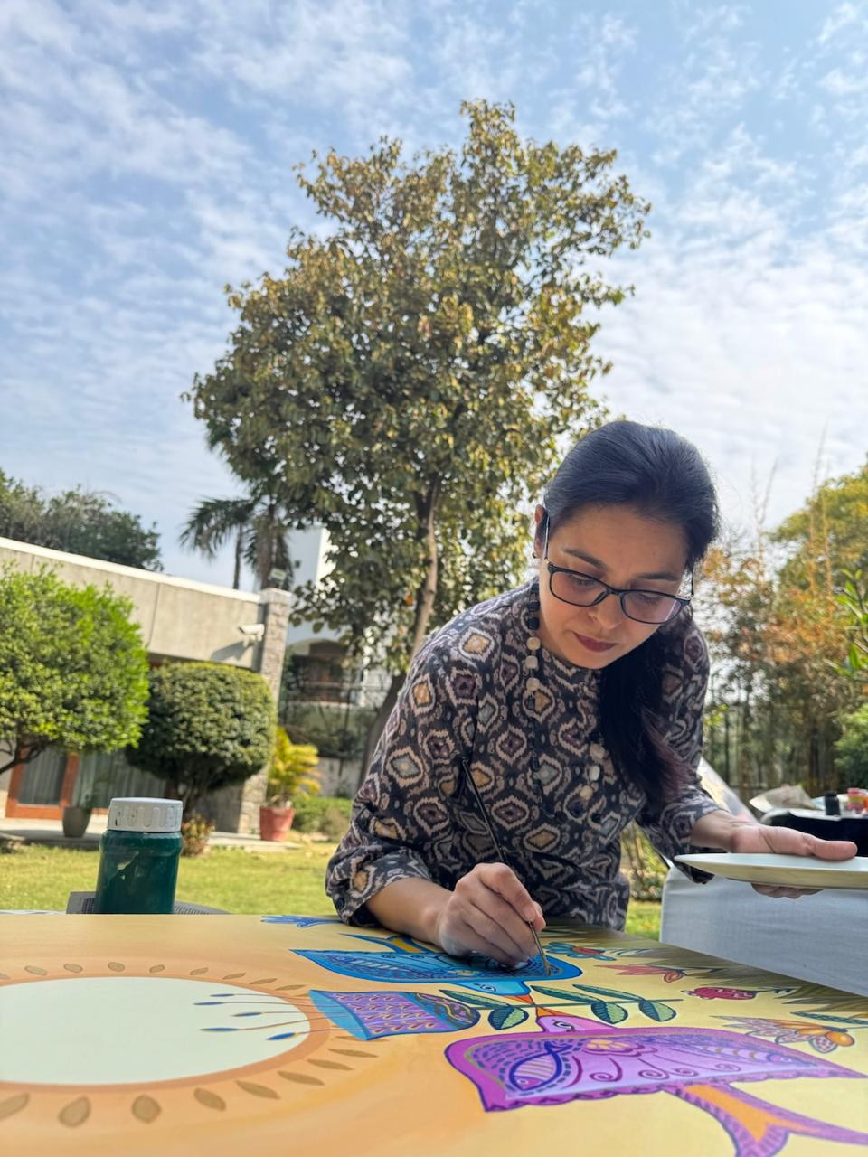 Pavit painting a blue bird on a golden canvas outdoors, with a palette in her hand.